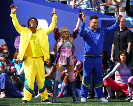 Ronaldinho, Madonna, and Ronaldo in colorful outfits raise their hands on a sports field with a crowd behind them